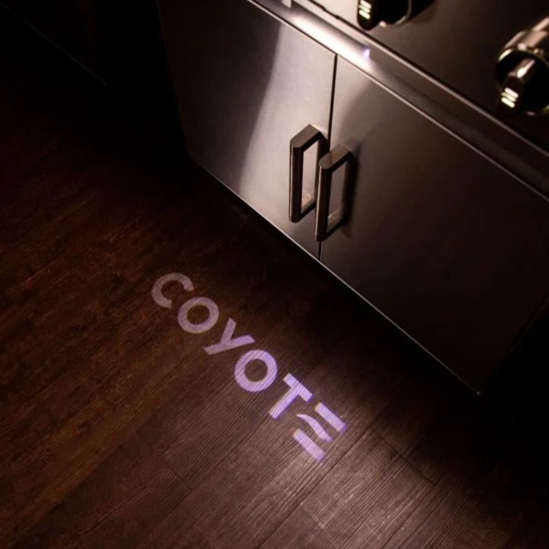 Coyote Grill Reviews – close-up of stainless steel grill cabinet with Coyote logo light projection, showcasing premium outdoor kitchen design.