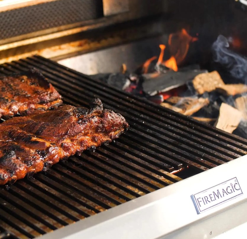 Close-up of ribs sizzling over open flame on a premium stainless steel Fire Magic grill—perfect for backyard chefs exploring the Fire Magic Grills FAQ and high-end outdoor cooking performance.