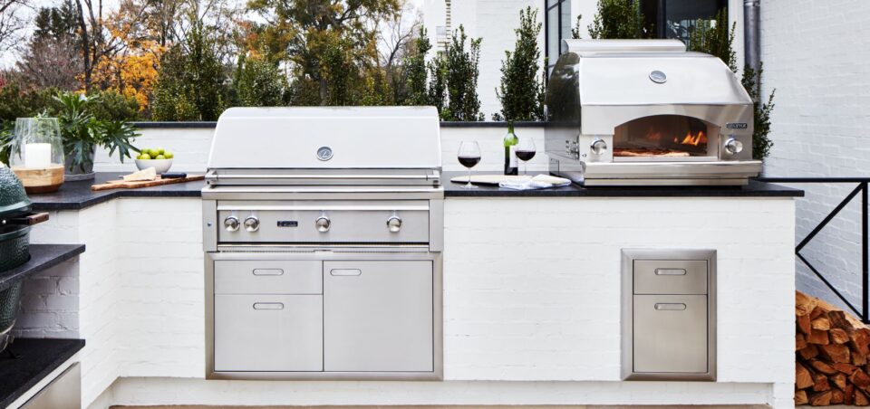 Lynx built-in grill and outdoor pizza oven installed in a modern outdoor kitchen island with stainless steel storage drawers and black stone countertops.