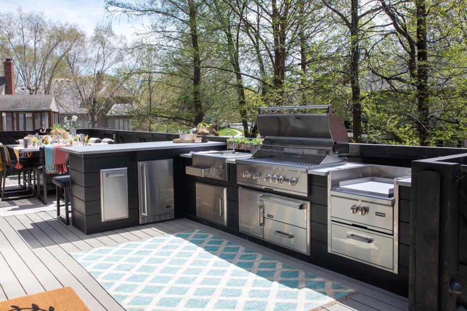 Modern outdoor kitchen featuring built-in Coyote grill, side burner, refrigerator, and storage drawers on a deck surrounded by trees