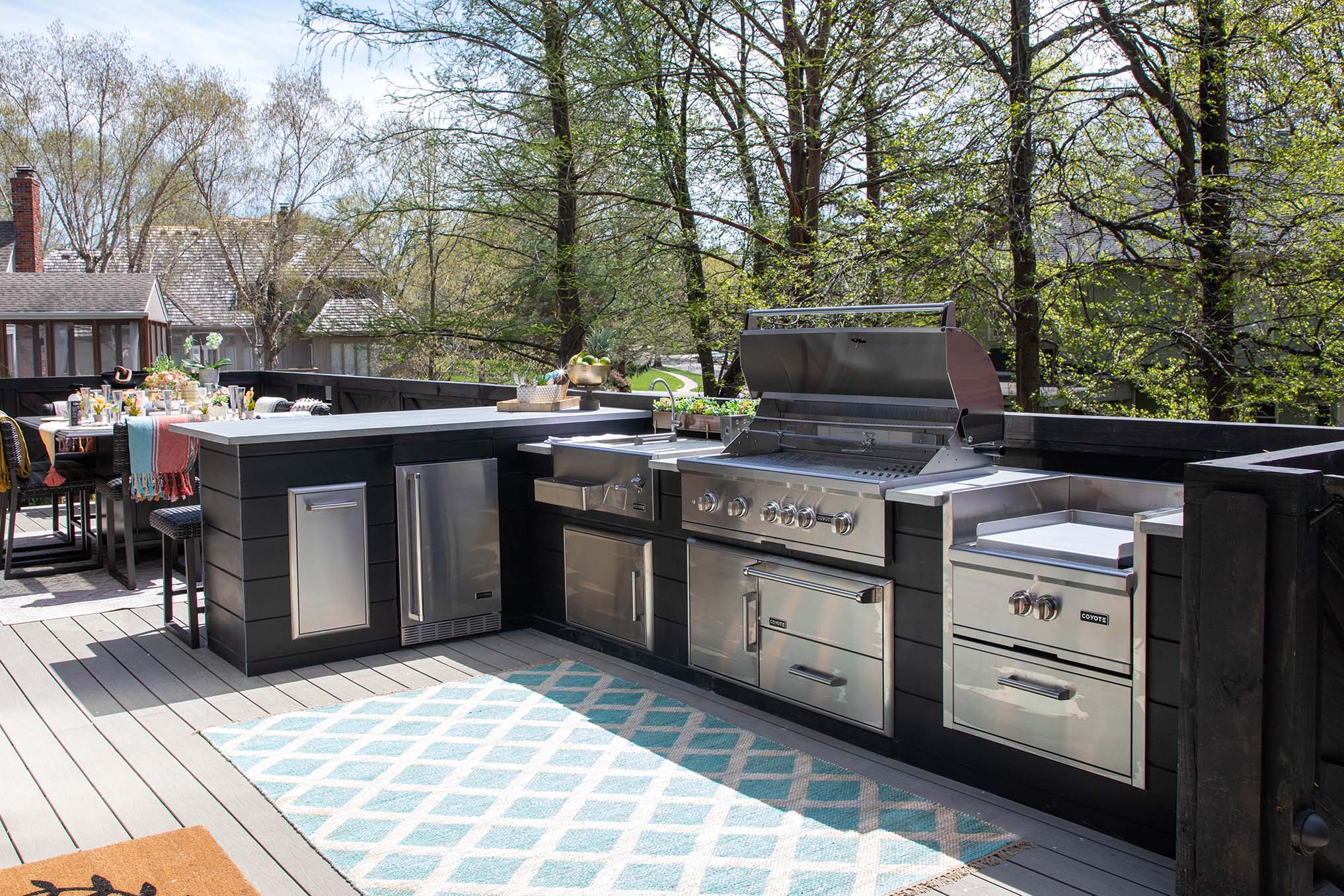 Modern outdoor kitchen featuring built-in Coyote grill, side burner, refrigerator, and storage drawers on a deck surrounded by trees