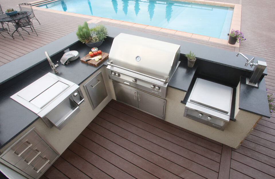 High-angle view of an Alfresco outdoor kitchen island featuring built-in luxury grill, sink, storage drawers, and accessories beside a backyard pool